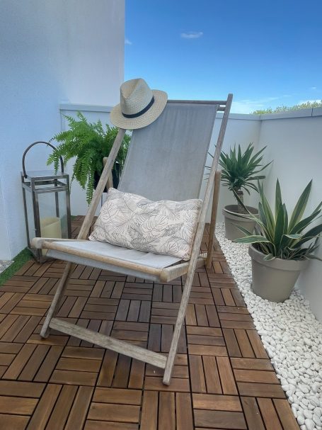 Chaise pliante avec coussin et chapeau, entourée de plantes et d'une lanterne sur un balcon.